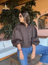 Camisa de color marrón oscuro con cuello clásico y mangas abullonadas con puños elásticos. Presenta un corte holgado y cierre frontal con botones.