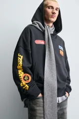 Sudadera tipo hoodie de corte relajado, color negro, con capucha y bolsillo frontal tipo canguro. Presenta parches decorativos en el pecho y estampados en color amarillo en la manga derecha. Acabados en rib en puños y ruedo.
