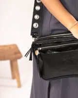 Bandolera negra de charol con doble compartimento, bolsillo frontal, correa regulable con ojalillos y detalles metálicos plateados.