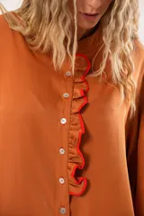 Camisa color naranja óxido con cuello mao, mangas largas abullonadas y cierre frontal con botones. Presenta un volante vertical con borde rojo en el frente.