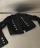 Campera estilo militar de color negro, con cuello mao y cierre frontal. Presenta múltiples botones metálicos plateados dispuestos en filas horizontales y detalles de charreteras en los hombros. Las mangas cuentan con puños decorados con botones a juego.