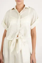 Camisa crop fluida de manga corta color crema con cuello camisero, botonadura frontal oculta y detalle de nudo en el frente.