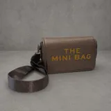 Cartera tipo bandolera de tamaño pequeño, confeccionada en material texturizado color bronce metalizado. Presenta el texto 'THE MINI BAG' estampado en letras doradas en el frente y cuenta con una correa de cinta ajustable al tono.