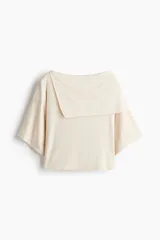 Top corto confeccionado en punto elástico de viscosa, color beige, con escote bote, hombros caídos y mangas cortas amplias. Presenta una sección superior superpuesta con un diseño asimétrico que crea un efecto capa.
