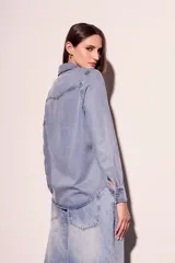 Camisa de denim celeste con cuello clásico, cierre frontal con botones a presión, dos bolsillos con solapa abotonada en el pecho y mangas largas con puños abotonados.