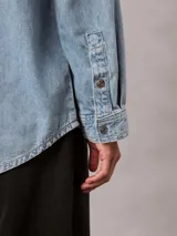 Camisa de denim de corte holgado, con cuello en punta, mangas largas y cierre frontal con botones. Presenta un diseño clásico con acabado lavado y monograma bordado en el pecho.