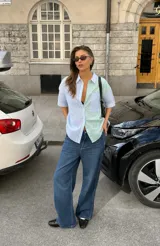 Camisa de diseño color-block a rayas, con un lado en tonos celeste y blanco y el otro en verde claro y blanco. Presenta mangas cortas, cuello camisero, cierre frontal con botones y un bolsillo aplicado en el pecho.
