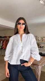 Camisa blanca de broderie con cuello redondo, cierre frontal con botones de madera y mangas largas abullonadas con puños elásticos.