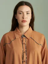 Camisa de algodón color tostado, con corte básico y caída suelta. Presenta cuello camisero, cierre frontal con botones y detalles de vivos con patrón geométrico en la botonera, canesú y hombros.