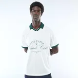 Camiseta tipo polo de manga corta, color blanco con cuello y ribetes en las mangas de color verde oscuro. Presenta un diseño gráfico estampado en el pecho con inscripciones y detalles en tono verde.