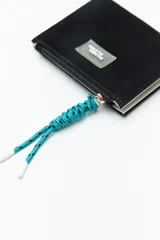 Billetera compacta de símil cuero color celeste claro, con cierre superior y un tirador de cordón trenzado multicolor. Posee un parche frontal con el logo de la marca.