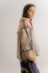 Chaqueta de paño color beige con costuras decorativas de crochet en colores vibrantes como rojo, azul, verde y amarillo. Presenta un diseño de corte holgado, cuello camisero y mangas largas.