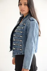 Chaqueta de estilo militar confeccionada en denim negro con efecto lavado. Presenta un diseño estructurado con cierre frontal de cremallera, cuello mao y múltiples botones metálicos decorativos dispuestos en filas horizontales. Incluye detalles de charreteras en los hombros y puños con botones.