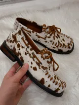 Zapato tipo mocasín de cuero con pelo estampado con diseño de manchas de vaca en tonos marrón y blanco. Presenta cordones frontales, costuras visibles en el borde y suela plana.