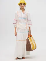 Falda larga blanca confeccionada en broderie de algodón con diseño de paneles escalonados, detalles de galones con transparencias en color azul y naranja, y pretina elástica con cordón ajustable.