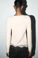 Top de manga larga en tejido suave color gris, con cuello redondo y bajo asimétrico decorado con encaje negro a tono. Presenta detalles de encaje negro en los puños.