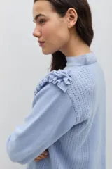 Sweater de lana color celeste, con cuello redondo, mangas largas y volados decorativos en los hombros. Presenta bordados florales artesanales en el frente y detalles de tejido calado vertical.