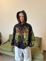 Hoodie oversize de color negro con estampado gráfico en pecho y mangas en tonos amarillos y verdes. Presenta un forro interior de capucha en color rojo con textura tejida y logo bordado en la manga.