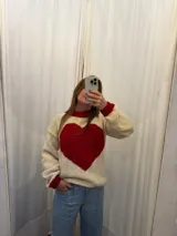 Buzo tejido a mano en lana merino, de color crema con un gran corazón rojo en el centro y detalles en rojo en el cuello y los puños. Presenta un diseño de manga larga y corte holgado.
