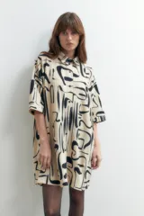 Vestido plisado color beige con estampado abstracto en negro, corte recto, cuello camisero y mangas cortas.