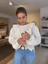 Blusa de tela liviana color crema con estampado floral pequeño en tonos rojizos. Presenta un diseño con volados en el frente y hombros, mangas largas abullonadas con puños elásticos y cuello redondo.