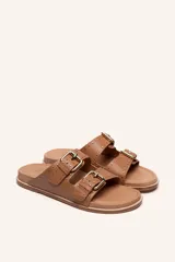 Sandalias de cuero color negro con dos tiras anchas sobre el empeine, cada una con hebillas metálicas ajustables y detalles de tachas plateadas. La suela es de cuero con un borde de color madera.