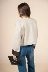 Chaqueta corta color beige, con cuello redondo, cierre frontal con botones dorados, bolsillos de parche y puños en contraste color marrón.