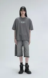 Remera de algodón premium color gris concreto, con corte oversize, hombros caídos y cuello redondo. Presenta un estampado frontal con las siglas "KM" y el texto "#KILOMBITOMAL" debajo en color celeste pálido.