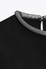 Remera negra de corte recto y holgado, con cuello redondo decorado con apliques de brillos y mangas cortas. Presenta una abertura en la espalda con cierre de botón oculto.