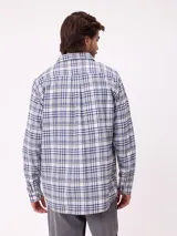 Camisa de manga larga con estampado de cuadros en tonos blanco, gris y azul. Presenta cuello clásico, cierre frontal con botones y un pequeño parche con logo en el pecho.