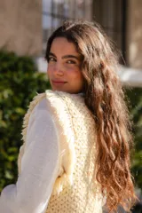 Chaleco de crochet tejido en punto cesta color crema, con diseño sin mangas y detalle de flecos en los hombros. Presenta un cierre frontal con alfiler de gancho metálico.