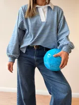 Buzo tipo chomba de corte oversize con diseño a rayas horizontales celestes y blancas. Presenta cuello camisero blanco y un bordado en la espalda con la inscripción República Oriental del Uruguay.