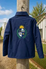 Campera impermeable de tela taslon color negro, con diseño de cuello alto y cierre frontal. Presenta un gran bordado en la espalda que incluye el escudo nacional uruguayo y el logo de la marca Bagual, además de un parche circular con la bandera de Uruguay en la manga.