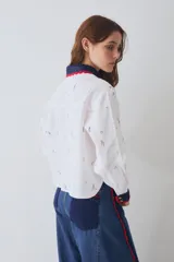 Camisa de lino blanca con estampado de motivos deportivos, cuello y puños tejidos a mano en crochet azul con borde rojo. Presenta cierre frontal con botones y un bolsillo en el pecho.