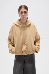 Campera de suede color beige con capucha, cierre frontal completo y mangas largas. Presenta un diseño holgado con bolsillos laterales y detalles de frunces en el ruedo.