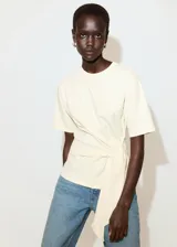 Camiseta color crudo de manga corta y cuello redondo, con diseño cruzado que forma un lazo lateral para anudar.