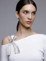 Sweater blanco de punto con escote asimétrico y manga larga. Presenta un broche con forma de moño con apliques de strass en el hombro.
