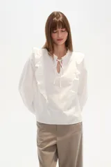 Blusa blanca de algodón con volados frontales, cuello redondo con lazo ajustable y mangas largas abullonadas con puños elásticos.