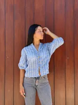 Camisa de corte recto y calce holgado, confeccionada en tela liviana color celeste con estampado de rayas verticales blancas con detalles de hojas. Presenta cuello en V, cierre frontal con botones de madera y mangas tres cuartos con terminación elástica.