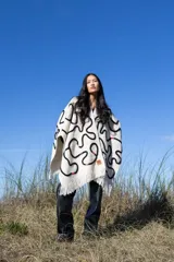 Ruana color crudo con diseño de líneas negras onduladas y detalles de cuentas de colores. Presenta flecos en el borde inferior.