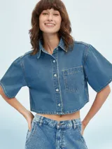 Camisa corta de denim azul claro, con cuello camisero, manga corta ancha y cierre frontal de botones blancos. Presenta un bolsillo de parche en el pecho izquierdo.