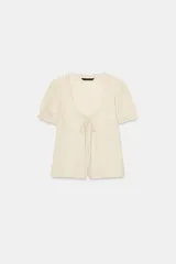 Camisa de gasa color crudo con escote en pico, manga corta abullonada con terminación en volante y cierre frontal con botones ocultos y lazada. Presenta delicados detalles de bordados a tono en toda la prenda.