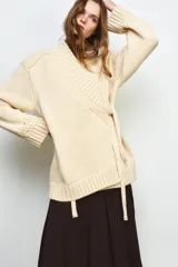 Chaqueta cruzada de punto color beige con manga larga, cuello smoking y cierre frontal con lazada. Presenta detalles de calados en los puños.