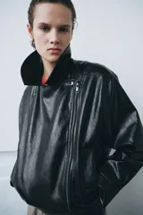 Chaqueta de estilo biker confeccionada en tejido efecto piel con acabado brillante. Presenta cuello con solapas, cierre frontal con cremallera y botones, bolsillos delanteros y bajo ajustable con trabillas y hebilla metálica.