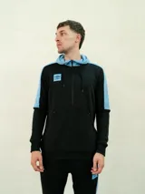 Canguro deportivo Umbro con capucha y cierre frontal parcial. Presenta un diseño en bloques de color negro y celeste, con logo de la marca estampado en el pecho y bolsillo frontal tipo canguro.