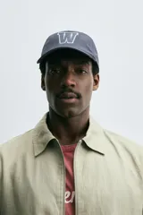 Gorra de seis paneles de tejido técnico color azul grisáceo, con visera curva y parche frontal con la letra "W" bordada en blanco y azul claro.