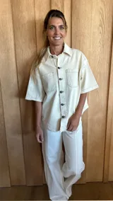 Sobrecamisa de denim soft color beige, con cuello clásico, cierre frontal de botones y mangas cortas. Presenta bolsillos frontales con solapa y costuras a la vista.
