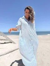 Vestido camisero midi de corte holgado, con estampado de rayas verticales celestes sobre fondo blanco. Presenta cuello camisero, cierre frontal con botones, mangas cortas con detalle de trabilla y bolsillos laterales.