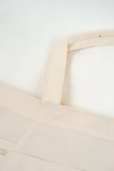Bolso tipo tote bag color beige con asas superiores de hombro, cordones ajustables a los lados con detalles metálicos y un pequeño anillo frontal.