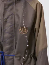 Campera tipo piloto con capucha, cierre frontal con botones y detalle de costuras en zigzag. Presenta cordones ajustables en la cintura y capucha, puños elásticos y logo bordado en el pecho.